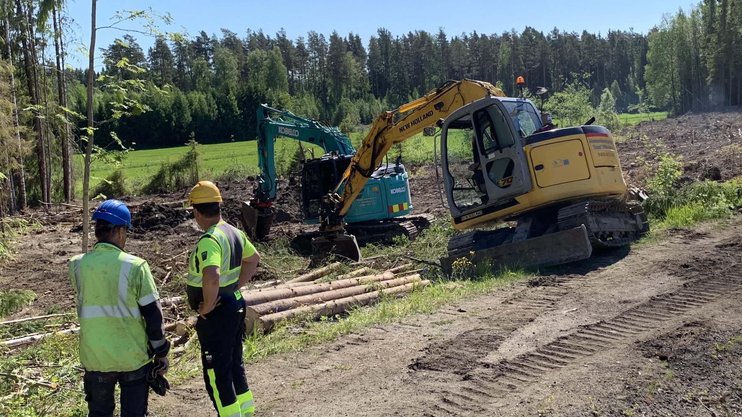 Kaksi henkilö etualalla ja kaksi kaivinkonetta maanrakennustöissä